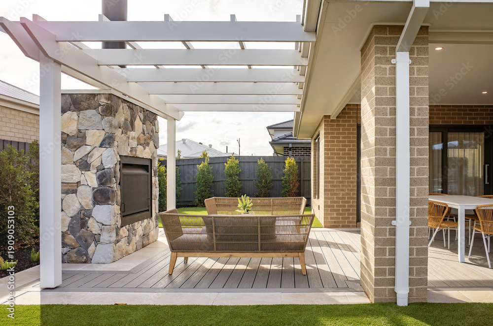 Fototapeta premium A covered outdoor living space with timber decking, pergola structure, and a stone feature wall fireplace of a contemporary suburban home. Outdoor landscaped backyard living in Australia.