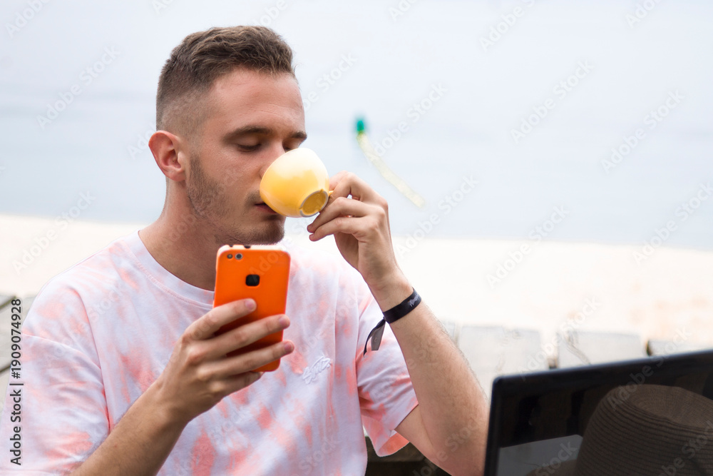 Fototapeta premium A young man checks his phone at a beach bar while drinking coffee.