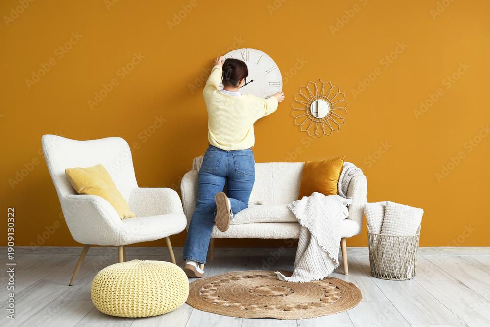 Fototapeta premium Young woman hanging big clock on orange wall in living room, back view