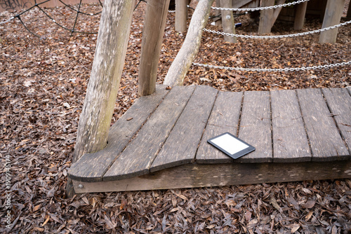 Ebook lying on a wooden playground structure outdoors. Everyday object in a casual setting, representing reading, learning, and leisure.