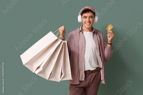 Young man in headphones with shopping bags and credit card on green background