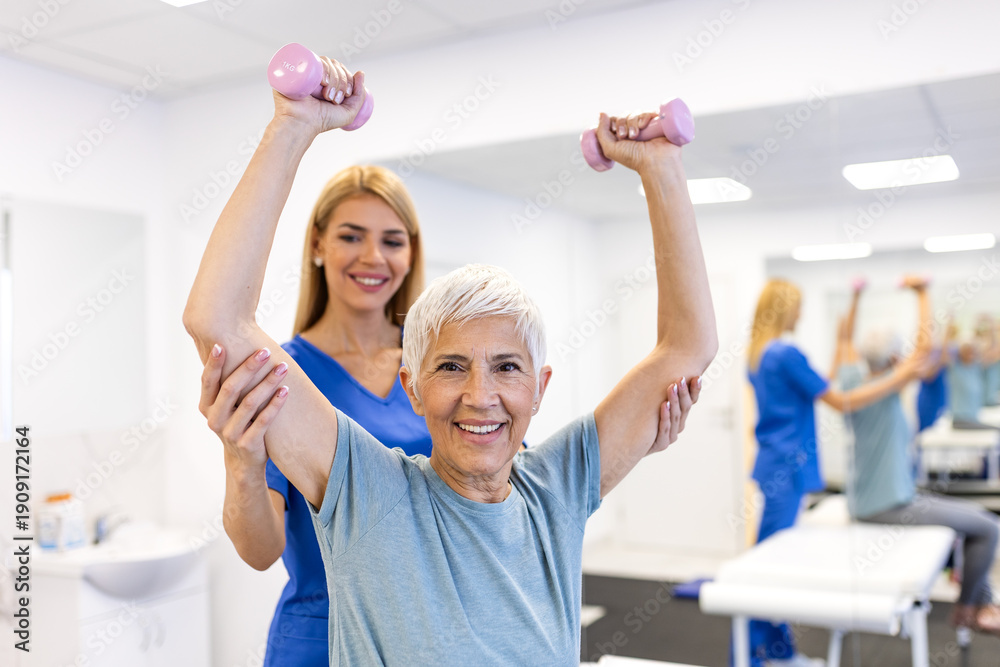 Obraz premium Happy senior woman lifting dumbbells with physiotherapist. Healthcare and medicine concept. Elderly patient doing strength training and rehabilitation exercises with female doctor in clinic office.
