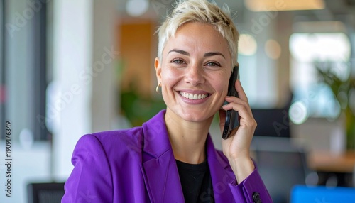 a smiling blonde woman with short hair wearing a purple blazer jacket making a telephone call in the office	