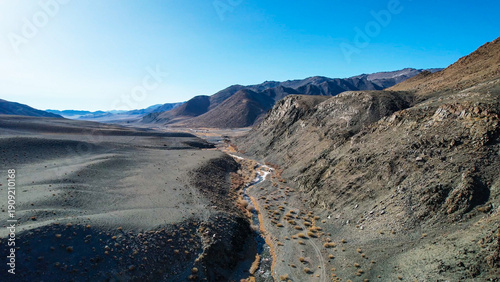 Sunlit Valley Winding Stream Through Arid Basin, Warm Tonal Contrasts Between Rocky Slopes And Blue Sky, Slender Watercourse Supporting Golden Grasses, Inviting Exploration And Contrast To Frozen
