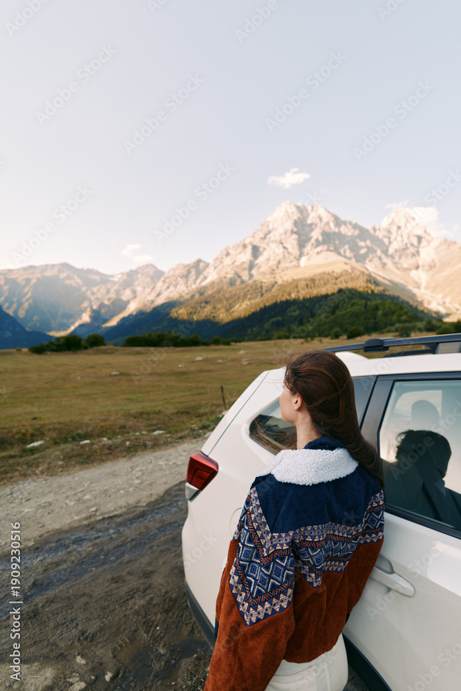 Fototapeta premium Woman in sweater beside car overlooking mountains and expansive landscape on a road trip, travel adventure and scenic journey outdoors at sunset with calm horizon and nature view.