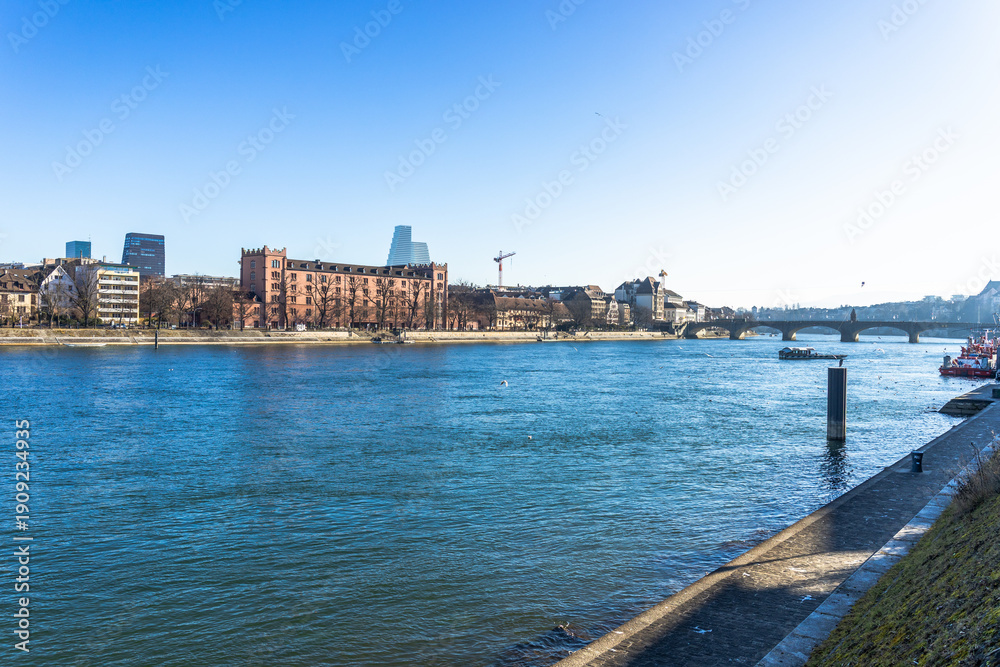 Obraz premium Scenic view of the Rhine river in Basel, Switzerland, featuring historic brick architecture on the riverbank under a bright blue sky.
