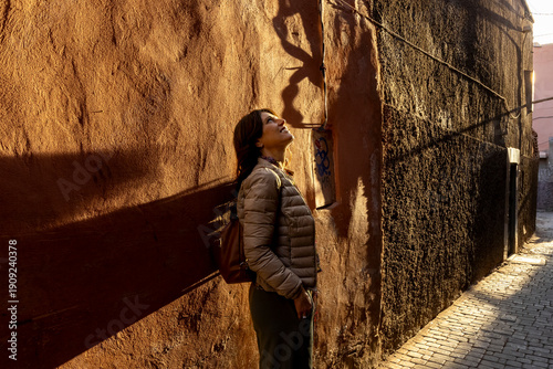 At Medina, a woman looks up at a shadow on a textured wall in a narrow alleyway, bathed in golden light. Jamaa el Fna,Marrakech,Unesco hertage,Morocco