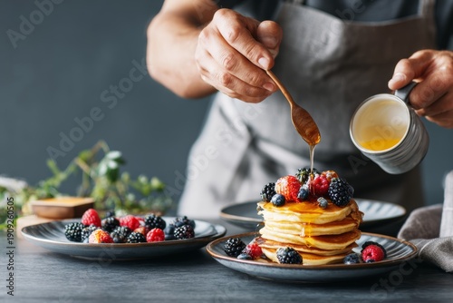 Professional chef in an apron pouring sweet honey syrup over a delicious stack of pancakes with fresh forest berries for a gourmet breakfast concept