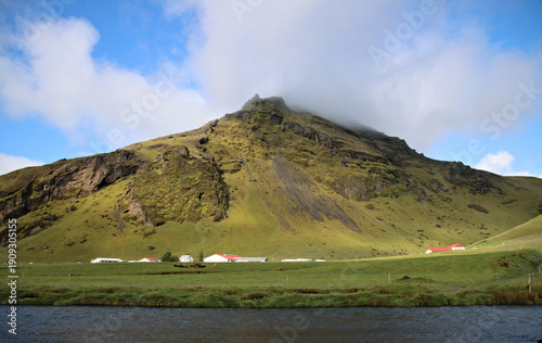 Rural farm buildings with red roofs beneath a steep green mountain with a mist-covered summit on the south coast of Iceland near Skogafoss, with a river flowing in the foreground.