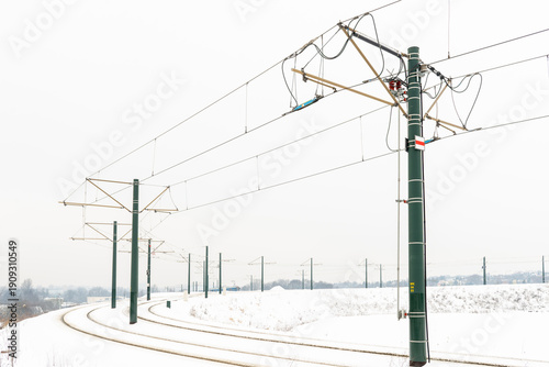 Curving railway track running through a snowy field with green catenary poles and overhead power lines. Minimal winter scenery highlights rail transport infrastructure, electricity supply, and cold-we