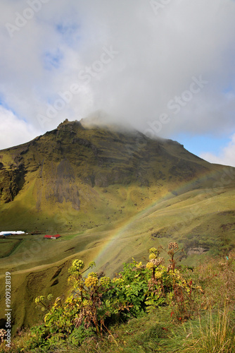 Rainbow forming across a green mountain slope with a cloud-covered summit near Skogafoss in southern Iceland, with rural buildings in the valley below.