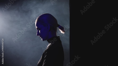 A young blonde woman with a short ponytail steps into a powerful side light. High-contrast video with her thoughtful profile illuminated, symbolizing the decision to stop being silent and embrace publ