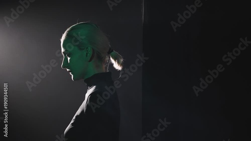 A young blonde woman with a short ponytail takes a step left into a dramatic beam of light. High-contrast video with side light highlighting her thoughtful profile. Concept of finding voice and facing