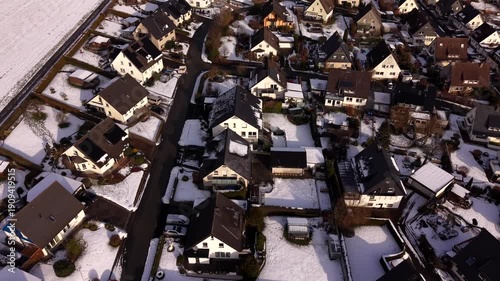 Aerial flyover snow-covered single family houses in German suburban neighborhood, showing quiet residential streets, winter conditions, rooftops, gardens and calm suburban environment. Top down shot.