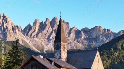 Aerial view of a quaint church nestled against the dramatic, jagged peaks of the Dolomites, a stunning contrast of architecture and nature, Santa Magdalena, Trentino-Alto Adige, Italy.