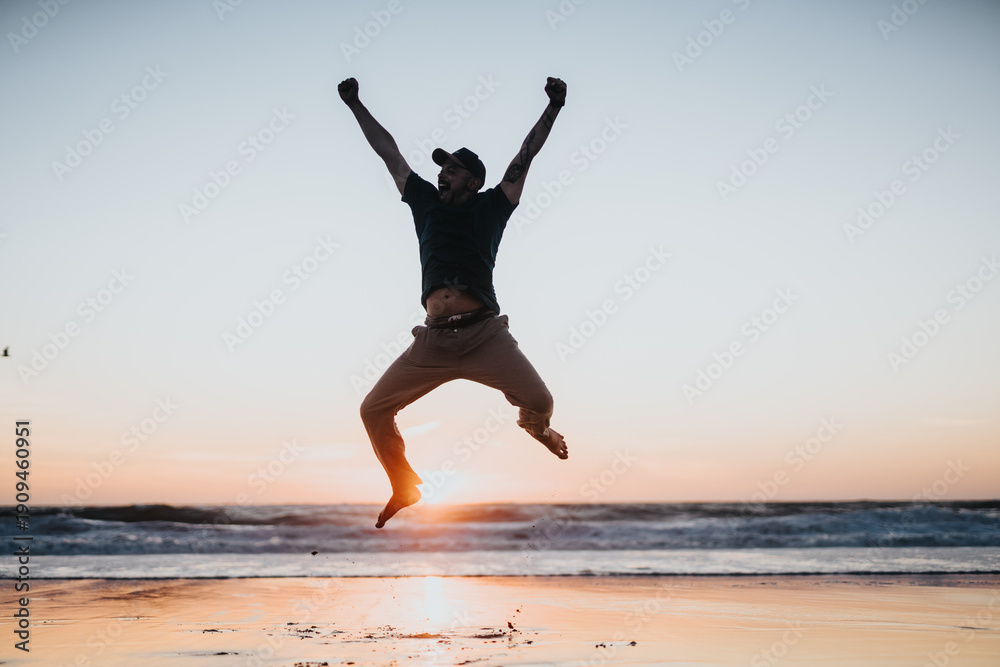 Obraz premium A man jumps mid-air on a sunlit beach at sunset, arms raised in victory. The scene conveys energy, freedom, and outdoor fitness against the calm ocean and warm sandy shore.