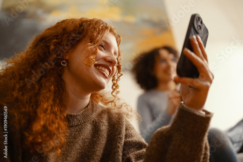 A young woman with curly red hair smiles as she holds up her phone, capturing a moment with a friend in the background.