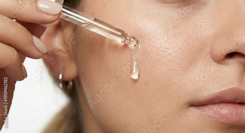 Close-up of a woman applying clear liquid serum with a glass dropper on her cheek, skincare routine and hydration concept