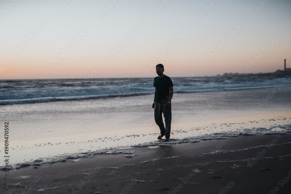 Naklejka premium A solitary man strolls the shoreline as gentle waves roll in during sunset, evoking calm, contemplation, and the serene mood of coastal life.