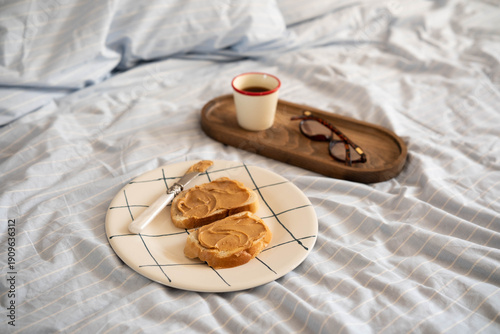 Obraz na płótnie A cozy breakfast in bed: two slices of toast with peanut butter on a patterned plate, a cup of coffee on a wooden tray, and a pair of glasses nearby