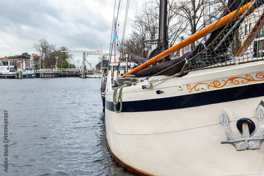 Fototapeta premium Traditional sailboat moored along canal Zwarte Water Netherlands near drawbridge village waterfront. Nautical detail rope anchor mast rigging under overcast sky creating calm maritime atmosphere