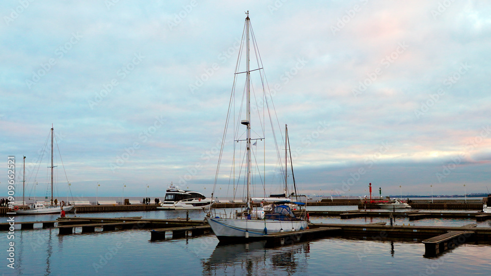 Fototapeta premium Sailboat anchored in harbor with cloudy sky and other boats during sunset