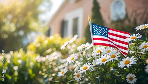 A small American flag waving in a garden full of white daisies in front of a house on a sunny day.