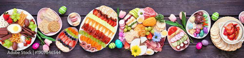 Spring or Easter grazing board theme table scene against a dark wood banner background. Overhead view. Assortment of sweets and cheese, meat and fruit appetizers.