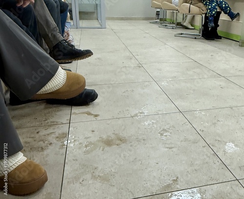 Patients Seated In Quiet Clinic. Silence And Routine Mark Quiet Clinic Waiting Area Atmosphere. Manicured Chairs And Faded Tiles Set Calm Tone For Patient Checkin Process