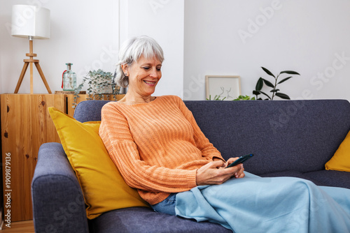 Konstfotografi Smiling senior woman using mobile phone relaxing on cozy sofa at home, browsing internet and connecting online in comfortable living room