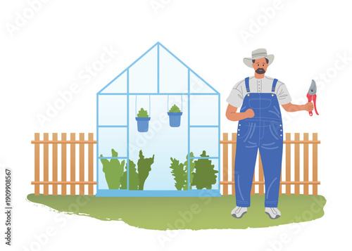 Male gardener in blue jumpsuit stands on lawn in front of greenhouse and holds pruner. Caring for fancy greenhouse plants.