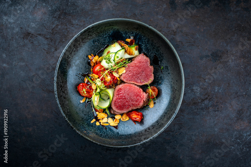 Fine dinining dry aged beef filet steak rare tataki style cut tagliata with sliced zucchini, tomato and almonds served as top view in a design bowl