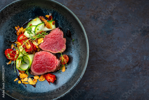 Fine dining dry aged beef filet steak rare tataki style cut tagliata with sliced zucchini, tomato and almonds served as top view in a design bowl with copy space right
