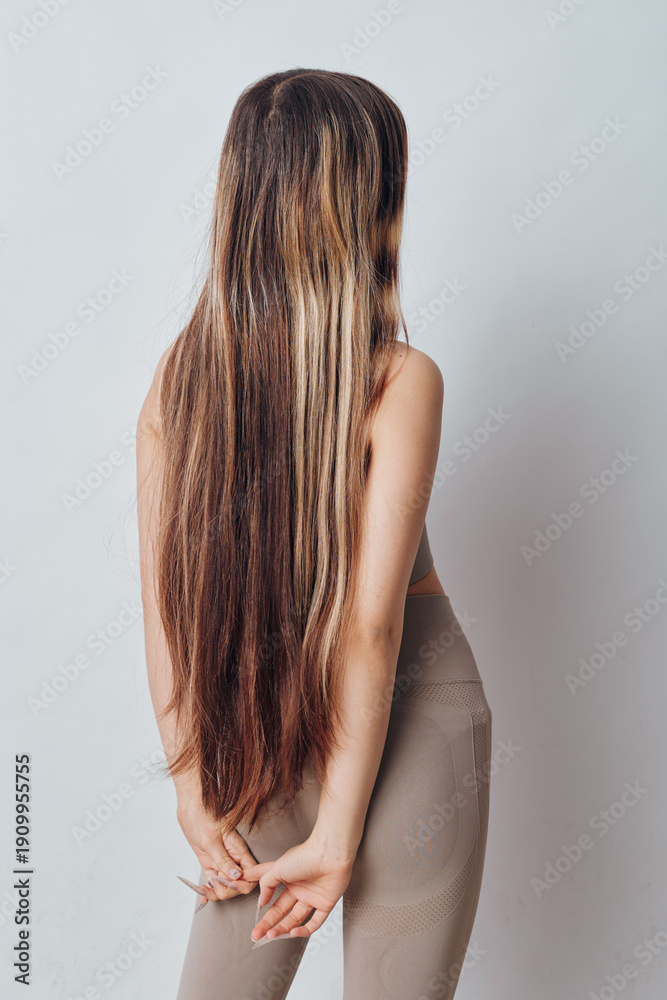 Obraz premium Woman with long straight hair standing with hands behind back wearing casual beige top and leggings in front of plain light background for beauty and style concept.