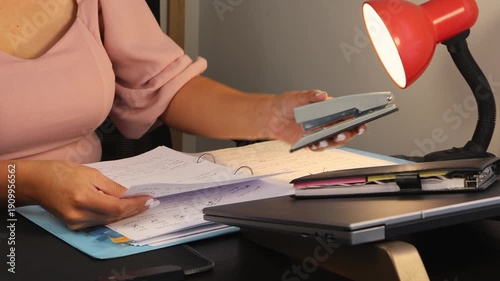 Close-up of a woman stapling papers at her desk. The image conveys office work, organization, productivity, and professional tasks in a modern workspace or home office environment.