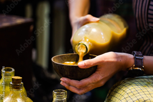 Jamu Herbal Medicine Pouring into Coconut Shell Bowl from Glass Bottle, Traditional Indonesian Healthy Beverage with Human Hand and Bamboo Basket, Organic Wellness Drink and Culinary Heritage