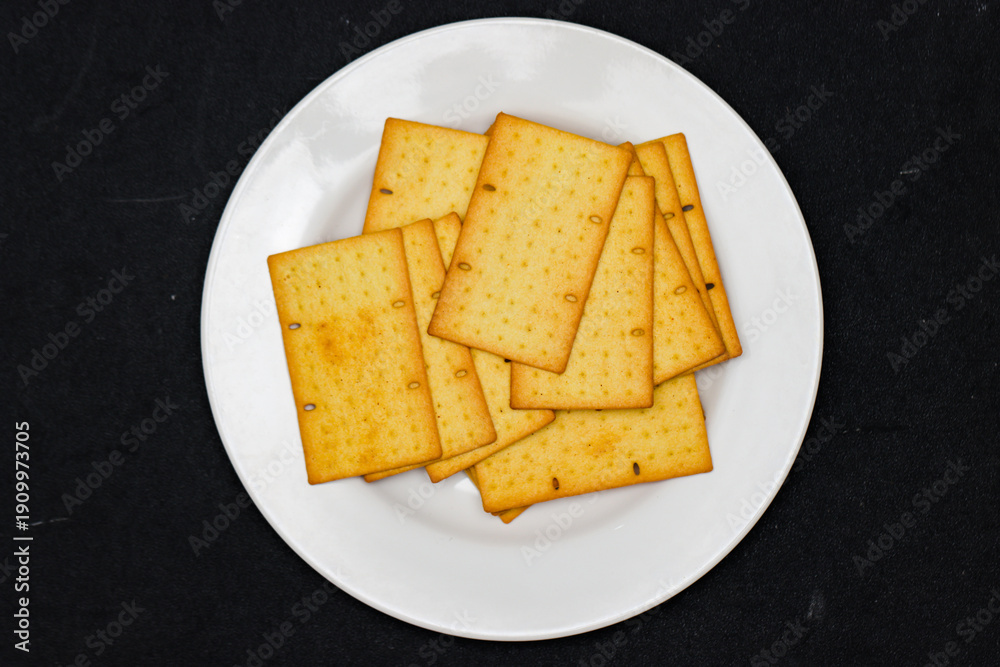 Fototapeta premium Crispy Wheat Crackers with Seeds on White Plate Top View, Savory Golden Brown Biscuits for Bakery and Culinary Branding, Healthy Crunchy Snack on Minimalist Black Background Flat Lay