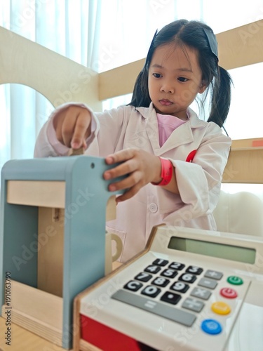 Картината върху платно Cute little Asian girl wearing a white coat playing pretend cashier with a wooden toy cash register in a bright playroom