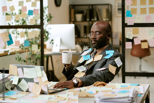 Konstfotografi Black man sitting at desk working on laptop surrounded by sticky notes holding c