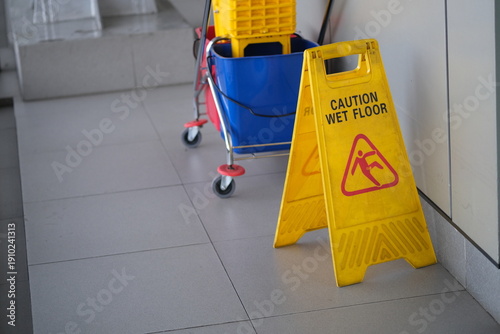 Yellow caution wet floor sign beside a janitor cleaning cart on tiled floor, highlighting safety warning, maintenance, and housekeeping in public spaces.