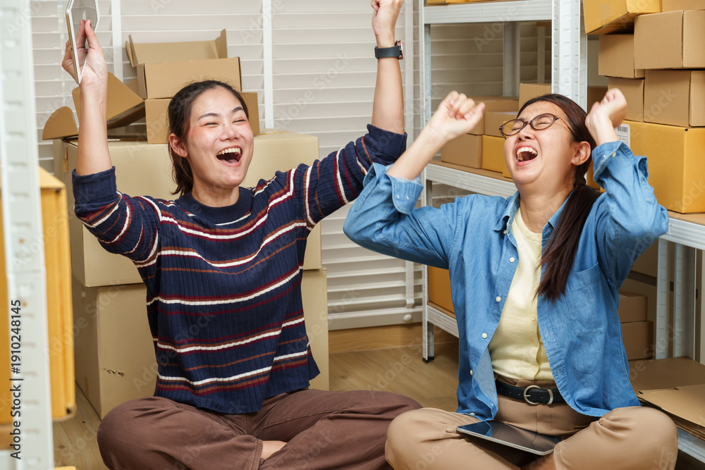 Obraz premium asian women partners raising arms and cheering after successful ecommerce order management celebrating online business achievement inside home warehouse with stacked parcel boxes
