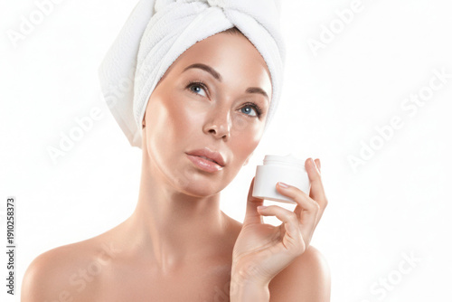 Beautiful Caucasian woman with white towel wrapped around her head. She holds cream jar in hand, isolated on white.