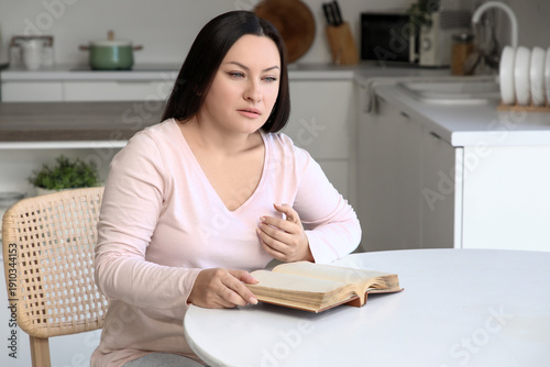 Young woman with book having heart attack at table in kitchen