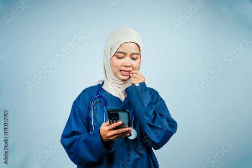 Anxious Asian Muslim nurse or doctor in navy scrubs and stethoscope biting her nail while looking at a smartphone. Concept of medical uncertainty, waiting for results, and stress.
