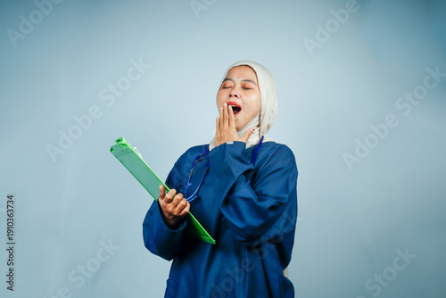Tired Muslim female nurse in hijab and blue scrubs yawning while holding a green clipboard. Exhausted healthcare professional showing signs of burnout during a long work shift.
