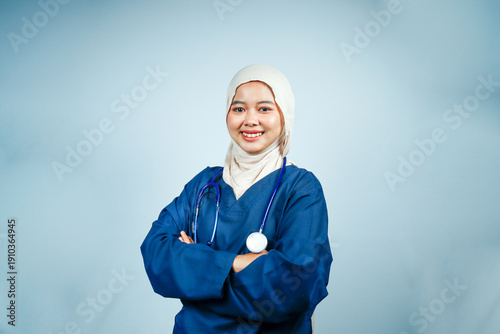 Confident Muslim female nurse in hijab and blue scrubs standing with her arms crossed. Friendly and professional healthcare worker posing for a medical staff portrait or bio.
