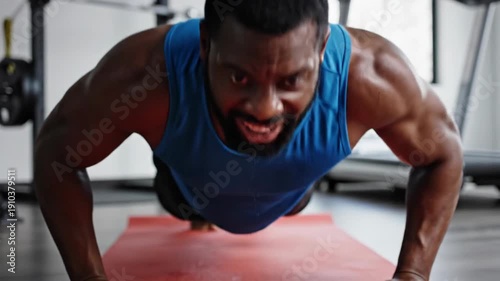 Muscular man in blue tank top struggles with push-ups on a red mat in a bright fitness space