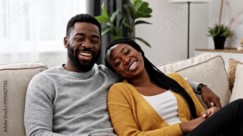 A Black couple laughs while sitting together on a couch in a cozy, relaxed living room setting