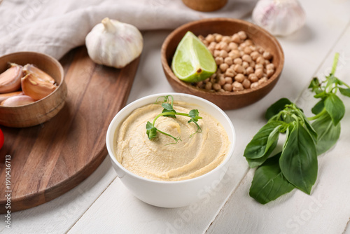 Fotografie Bowl of tasty hummus with ingredients, board and napkin on white wooden backgrou