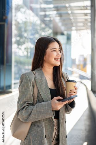 Happy asian woman office worker holding coffee cup and phone while standing sideway aside building.